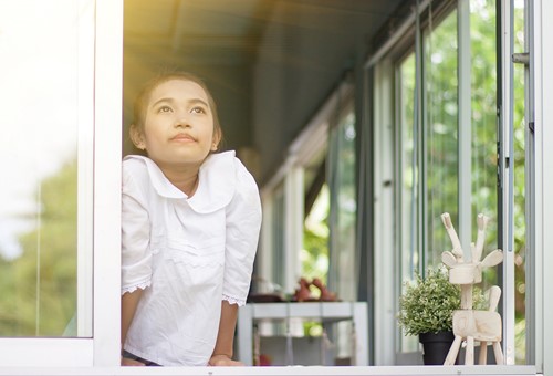 A young Asian girl looks out the window of her house at nature with a bright light in the sky.